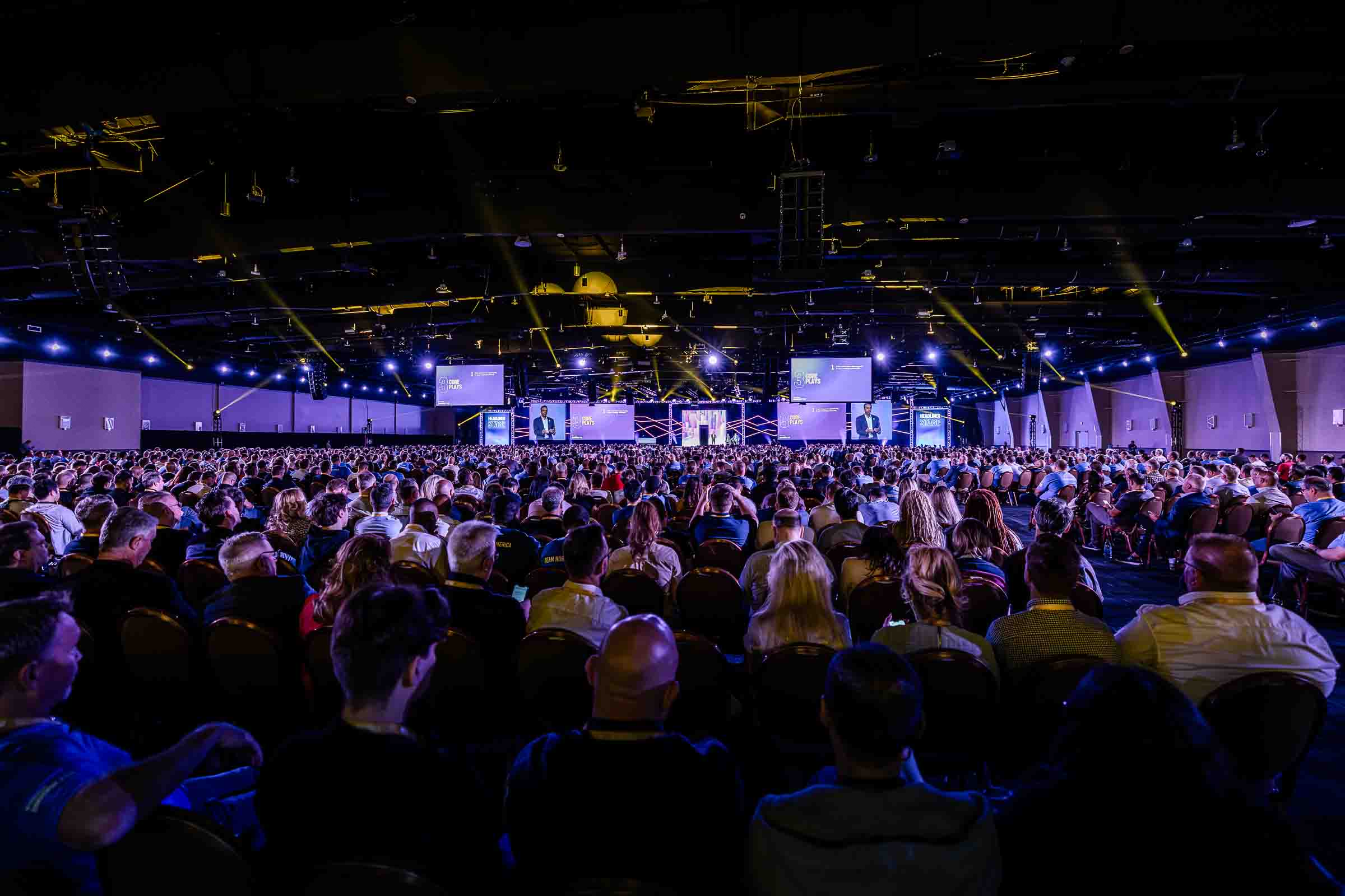 Massive corporate keynote audience with dramatic blue lighting and golden spotlight beams directed at stage with video screens